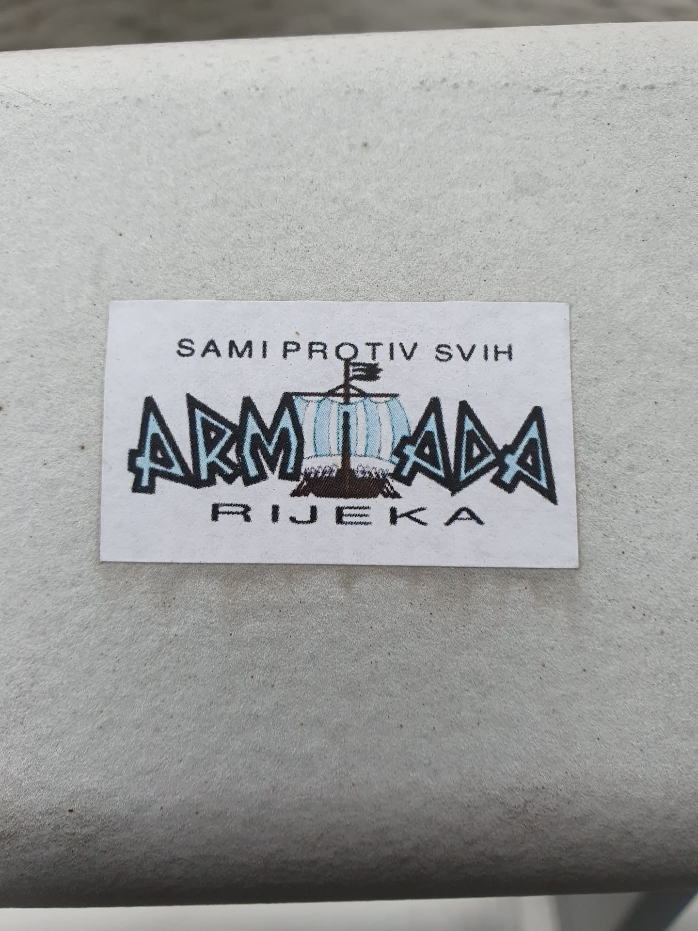 A sticker featuring the Armada Rijeka logo, a stylized ship with text above 'SAMI PROTIV SVIH' which means 'alone against all' and below 'RIJEKA', which is the name of the city. The overall design is simple yet striking.