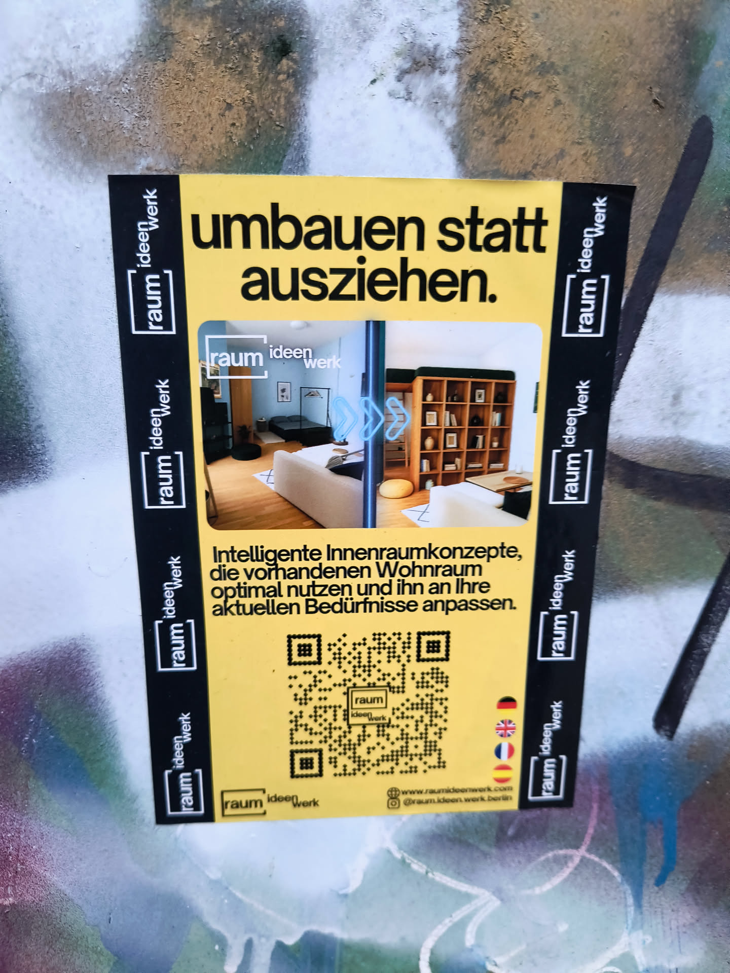 The sticker is predominantly yellow with a black border. The top of the sticker features bold black text that reads 'umbauen statt ausziehen.' (renovate instead of moving out.). Below this, there are two contrasting images of interior spaces: one showing a modern living room with a sofa and a bed, and another displaying a well-organized bookshelf unit. A QR code is prominently placed in the lower half of the sticker, surrounded by smaller images of flags (German, UK, Spanish). The black border on the left and right sides of the sticker is repeated vertically with the text 'raum ideen werk' in white, enclosed in a black rectangle.
