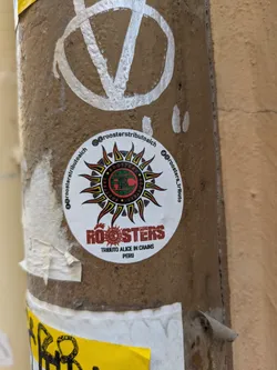 A round sticker on a pole featuring the logo for "Roosters Tributo Alice In Chains Peru". The logo consists of a sun-like burst of colored lines with the stylized text "AIC" in the middle and "ROOSTERS" in bold red letters below. The sticker has social media handles around the edge.