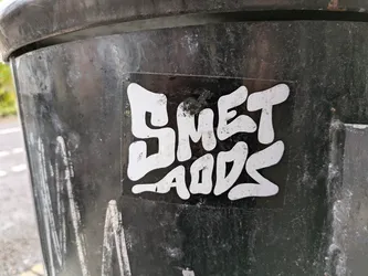 A white sticker on a black surface with the words SMET AODS written in a stylized, graffiti-like font. The letters are bold, slightly irregular, and appear hand-drawn or digitally created to mimic a hand-drawn style. The sticker shows some signs of wear.