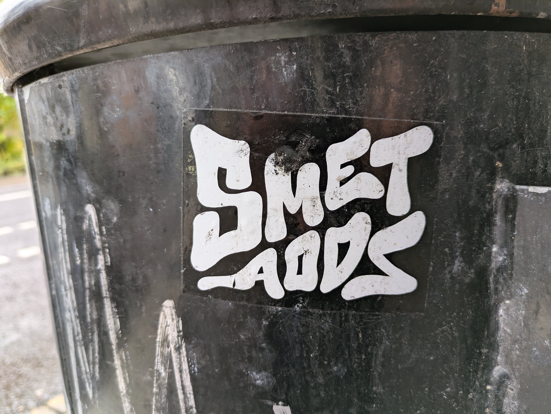 A white sticker on a black surface with the words SMET AODS written in a stylized, graffiti-like font. The letters are bold, slightly irregular, and appear hand-drawn or digitally created to mimic a hand-drawn style. The sticker shows some signs of wear.