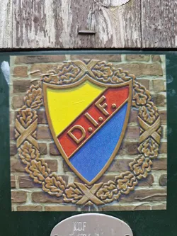 A sticker featuring an emblem with the letters D.I.F. in a shield surrounded by an oak leaf wreath. The shield is divided into yellow, red, and blue sections. The sticker appears to be affixed to a brick wall.