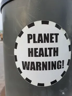 A round sticker with a black and white design.  It has a dashed black circle around the edge and bold black text on a white background that reads: PLANET HEALTH WARNING!