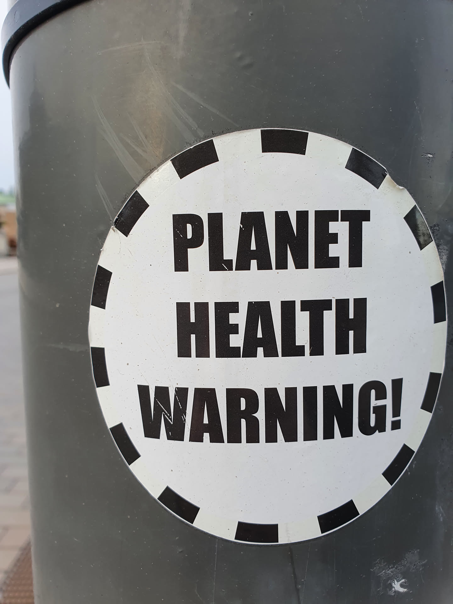 A round sticker with a black and white design.  It has a dashed black circle around the edge and bold black text on a white background that reads: PLANET HEALTH WARNING!