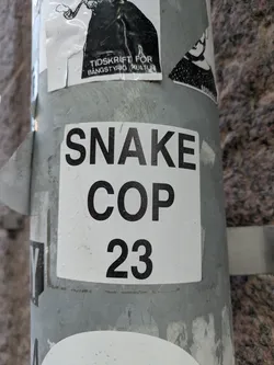 A close-up shot of a square sticker on a pole. The sticker is white with black text that reads "SNAKE COP 23".