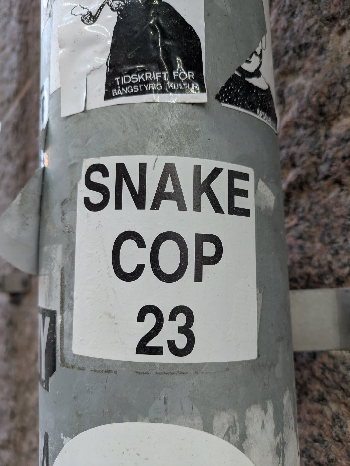 A close-up shot of a square sticker on a pole. The sticker is white with black text that reads "SNAKE COP 23".