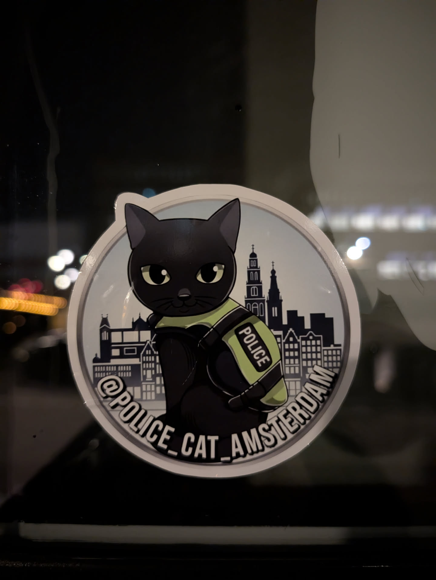 A circular sticker featuring a stylized black cat with large, light green eyes, wearing a bright lime green police vest with 'POLICE' written vertically in white. Behind the cat is a silhouette of the Amsterdam skyline, including iconic canal houses and church spires, rendered in shades of grey and white against a light blue background. The sticker has a white outer border, and along the bottom edge, in a curved arc, is the text '@POLICE_CAT_AMSTERDAM' in white letters with a black outline. The overall aesthetic is cartoonish and charming.