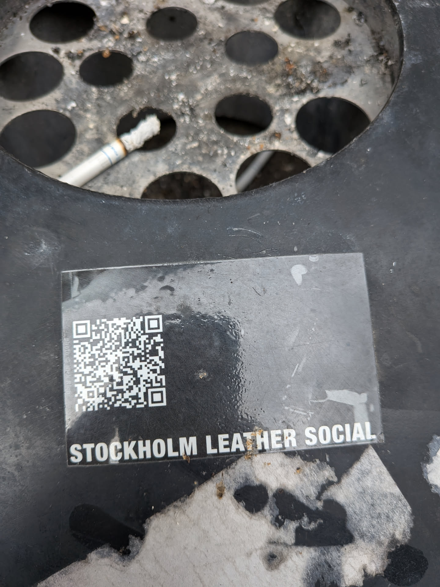 A rectangular sticker, predominantly black, featuring a QR code on the left side and white text on the right. The QR code is clearly defined with black and white squares. Below the QR code, the text 'STOCKHOLM LEATHER SOCIAL' is printed in bold, sans-serif white capital letters. The sticker appears to be slightly glossy and has some minor scuffs and what looks like a small water droplet or smudge on its surface.