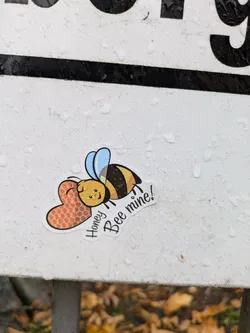 A cute bee sticker with a heart. The bee is yellow and black with blue wings. The heart is orange and has a honeycomb pattern. The text says "Honey Bee mine!"