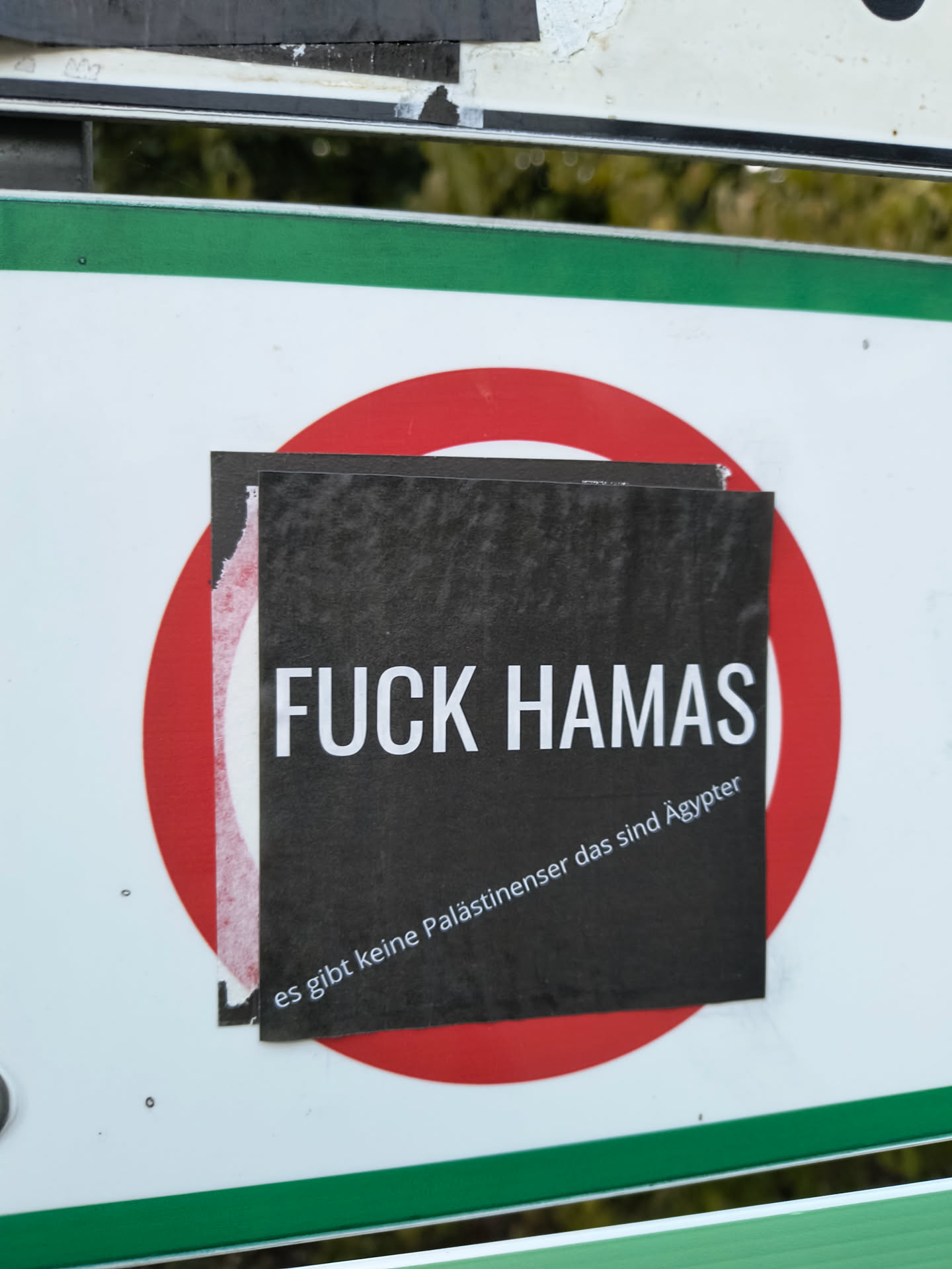 The sticker is black with white text. The top line reads 'FUCK HAMAS' in large, bold, sans-serif capital letters. Below this, in smaller, italicized text, it says 'es gibt keine Palästinenser das sind Ägypter'. The sticker appears to be slightly torn on the left edge, revealing a white backing and a red circular shape behind it. The overall impression is one of a printed paper sticker applied to a surface.