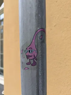 The sticker features a cartoon character with a rounded, tear-drop shaped pink body that tapers to a curved stalk at the top. It has small, stubby arms and legs, and appears to be wearing large, square sunglasses with a blue frame. The character also has prominent, red lips and a slight frown. Several small, pink, petal-like shapes are scattered around the character, some appearing to drip from its body. A circular element with text is visible within the character's stalk, and other small text is present on the sticker below and to the left of the character. The overall color palette is dominated by pink, with black outlines and blue accents for the sunglasses.