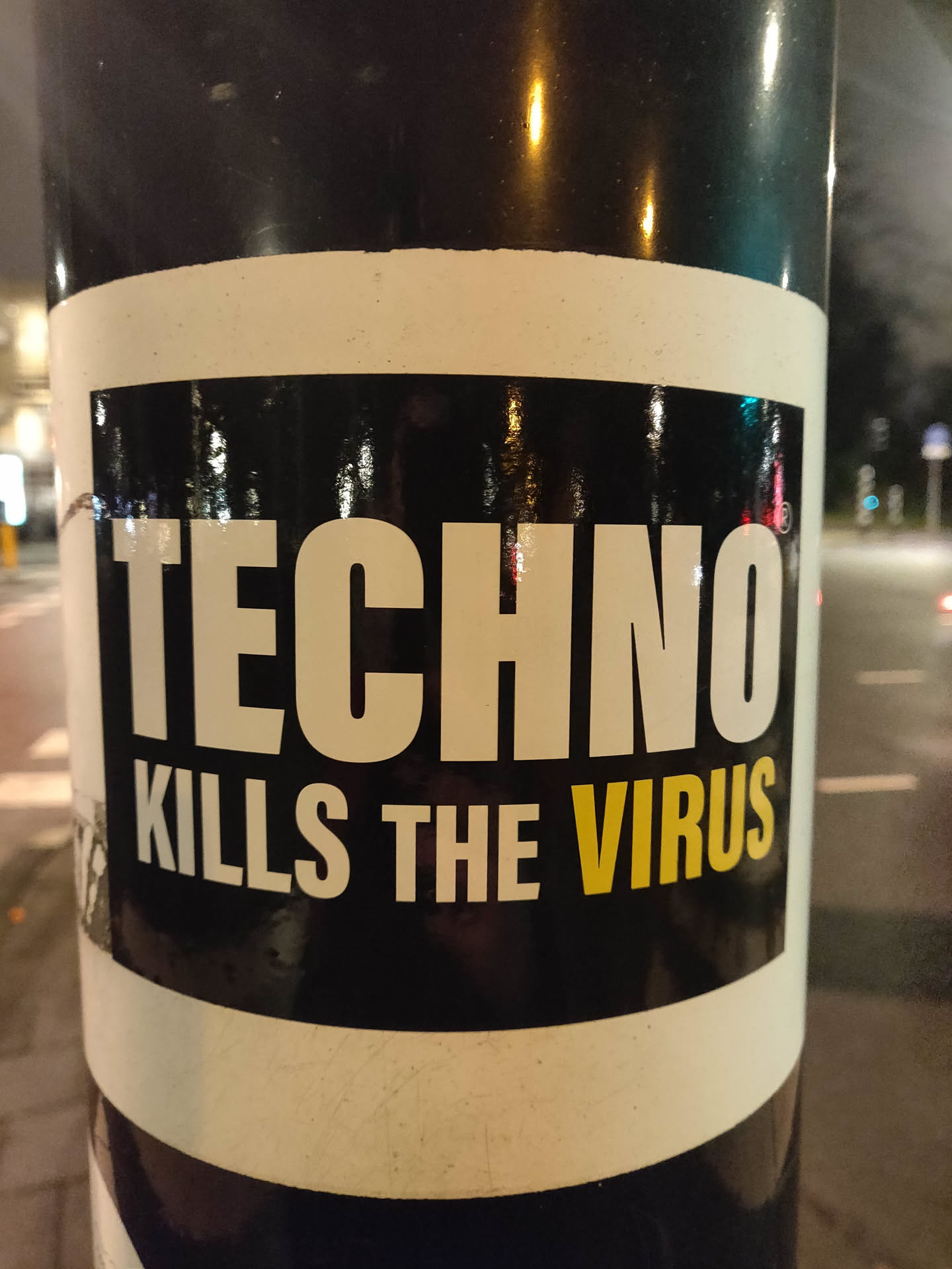 A black and white sticker with the text "TECHNO KILLS THE VIRUS" on a black background with a white border. The sticker is affixed to a dark-colored pole outdoors at night.