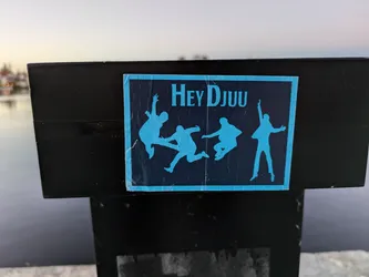 A sticker featuring the text 'Hey DJUU' and several stylized silhouettes of people jumping. The sticker is rectangular and blue with dark blue text and images, affixed to a black surface outdoors near water.