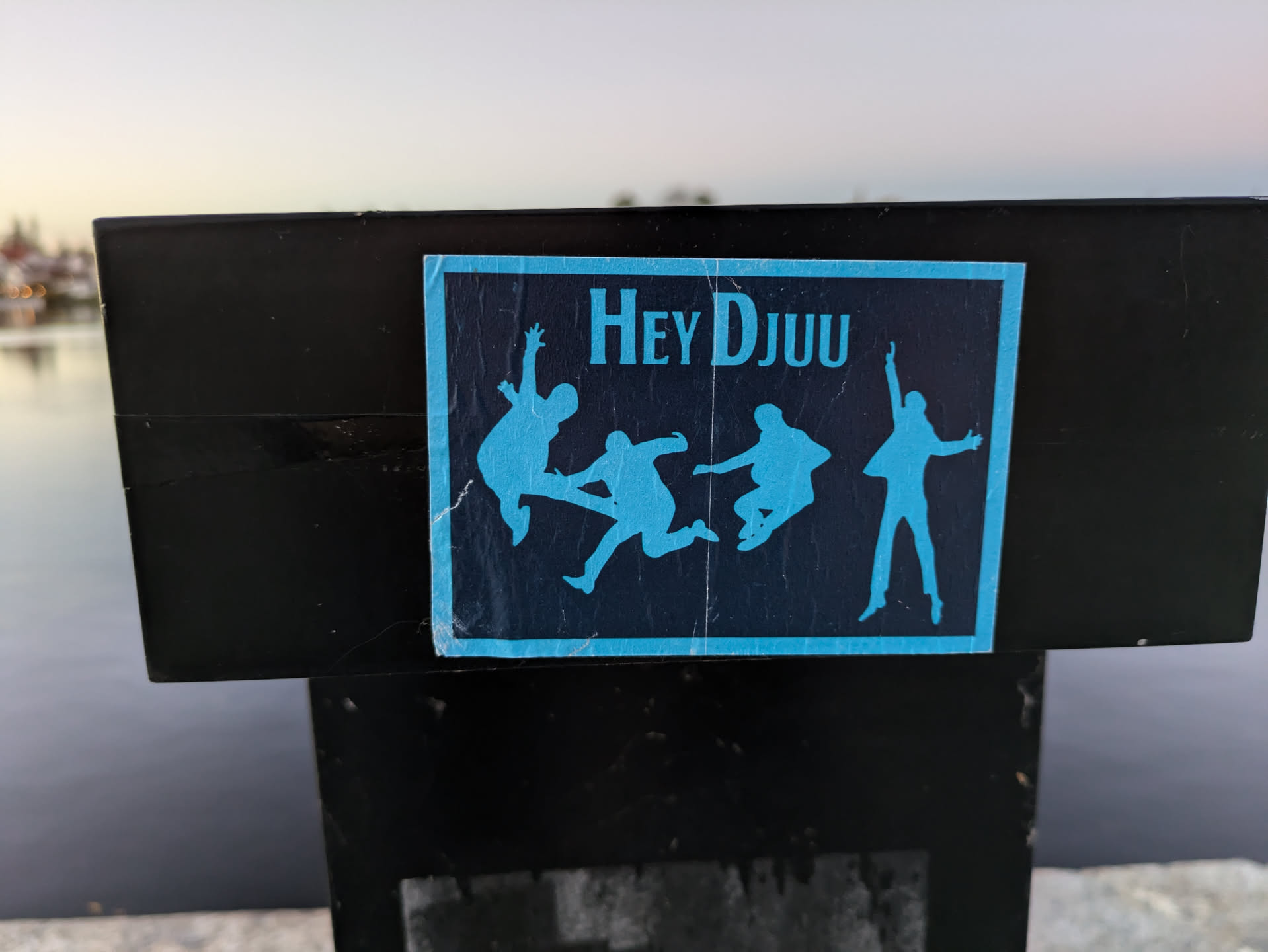 A sticker featuring the text 'Hey DJUU' and several stylized silhouettes of people jumping. The sticker is rectangular and blue with dark blue text and images, affixed to a black surface outdoors near water.