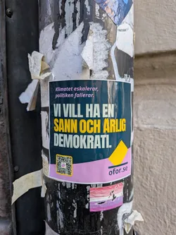 The sticker features a dark teal background with a prominent pink band across its lower section. At the top, the text 'Klimatet eskalerar, politiken fallerar.' (The climate escalates, politics fails.) is written in pink. The main message, 'VI VILL HA EN SANN OCH ÄRLIG DEMOKRAT!' (WE WANT A TRUE AND HONEST DEMOCRAT!), is displayed in bold, with 'VI VILL HA EN' and 'DEMOKRAT!' in white, and 'SANN OCH ÄRLIG' highlighted in yellow. On the left side of the pink band, there is a QR code with a yellow border and a small pink square in its center. To the right of the pink band, the website 'ofor.se' is printed in dark text, accompanied by a small yellow triangular graphic above it. The sticker is affixed to a weathered, black pole, surrounded by remnants of other torn papers.