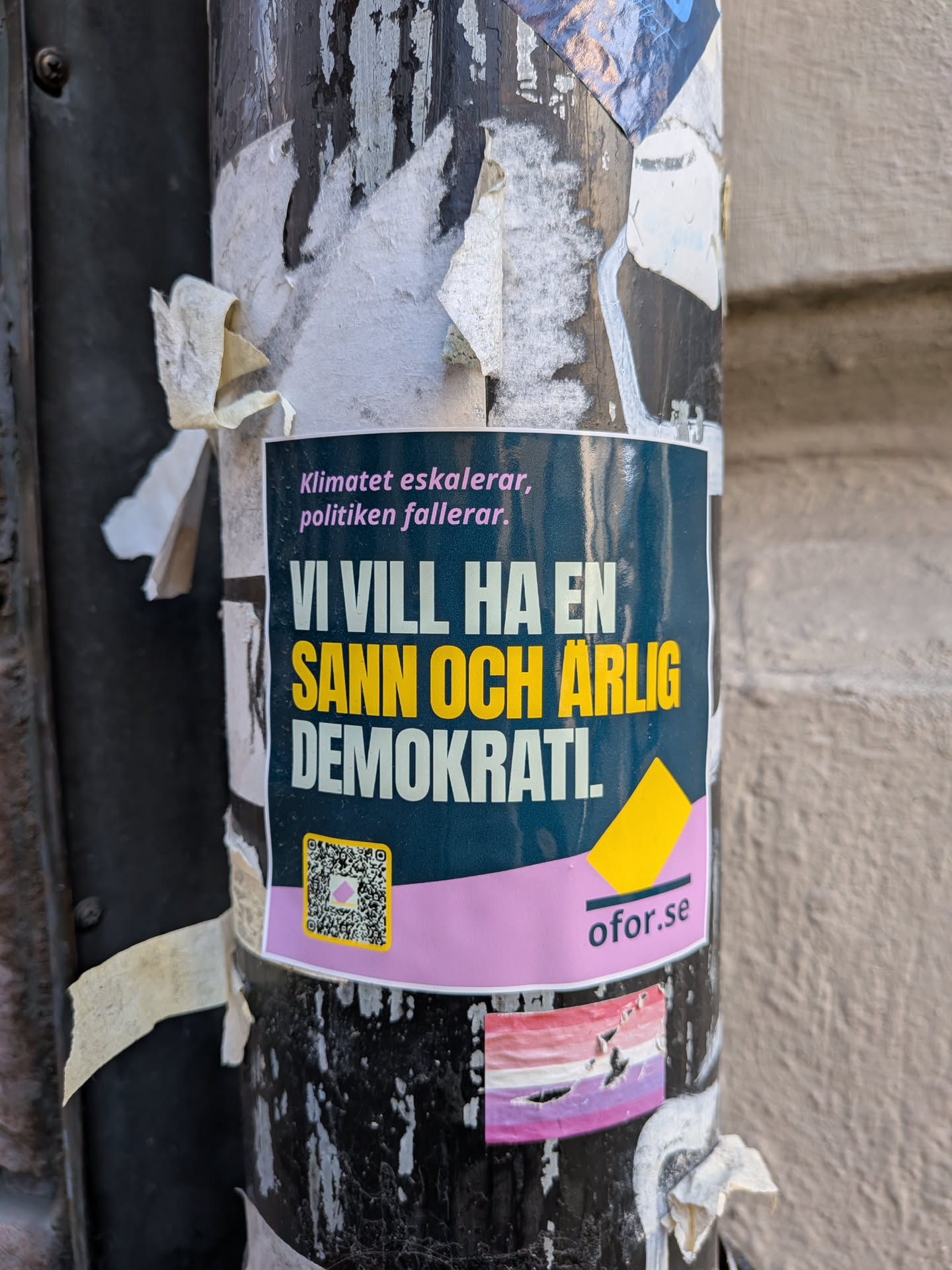 The sticker features a dark teal background with a prominent pink band across its lower section. At the top, the text 'Klimatet eskalerar, politiken fallerar.' (The climate escalates, politics fails.) is written in pink. The main message, 'VI VILL HA EN SANN OCH ÄRLIG DEMOKRAT!' (WE WANT A TRUE AND HONEST DEMOCRAT!), is displayed in bold, with 'VI VILL HA EN' and 'DEMOKRAT!' in white, and 'SANN OCH ÄRLIG' highlighted in yellow. On the left side of the pink band, there is a QR code with a yellow border and a small pink square in its center. To the right of the pink band, the website 'ofor.se' is printed in dark text, accompanied by a small yellow triangular graphic above it. The sticker is affixed to a weathered, black pole, surrounded by remnants of other torn papers.