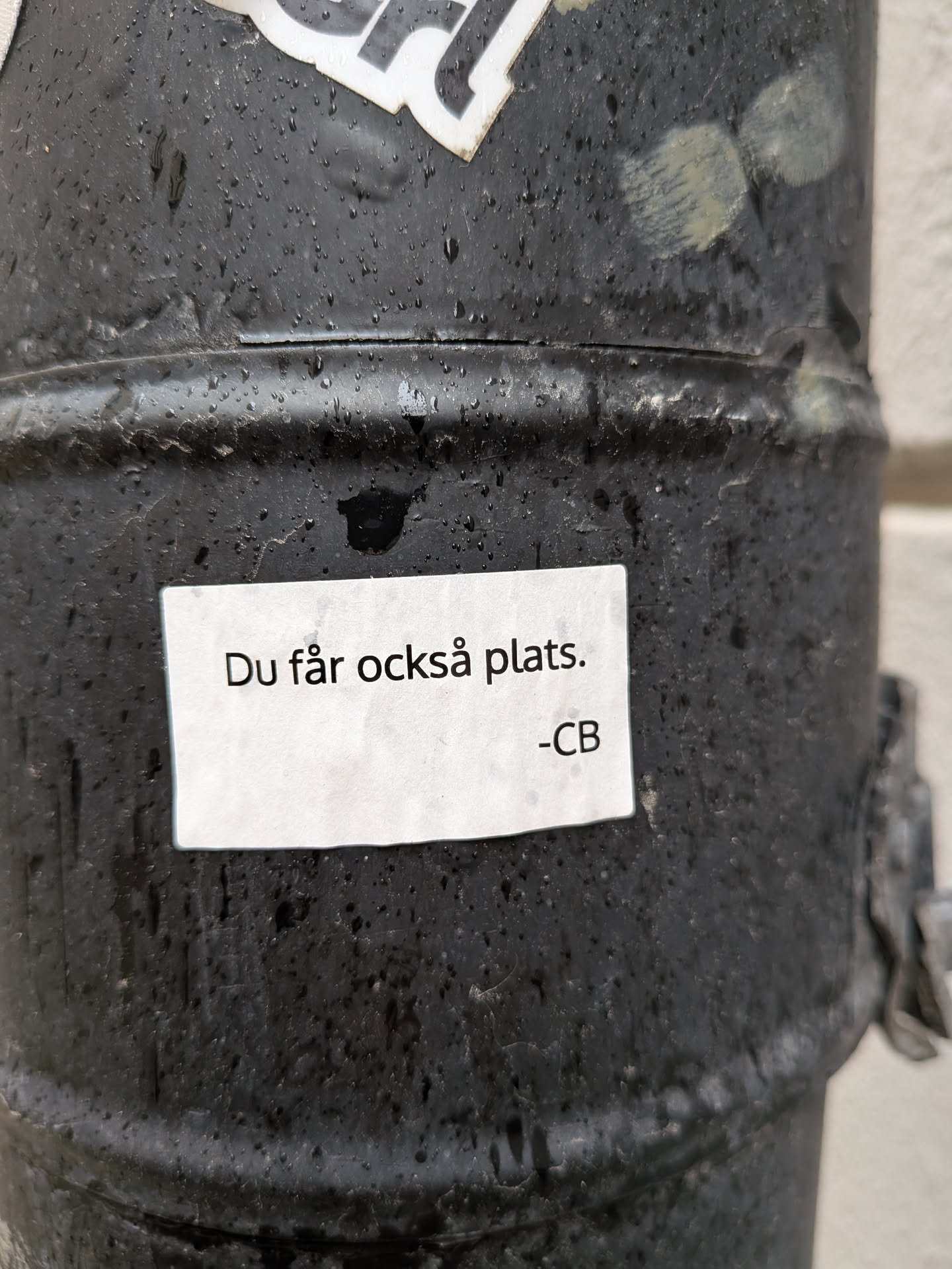A rectangular white sticker with black text, featuring the phrase 'Du får också plats.' (You also get a place.) and the initials '-CB' below it. The text is printed in a clean, sans-serif font. The sticker is slightly damp and adheres to a dark, textured, wet metal surface, possibly a drainpipe or pole. Its edges show minor wear, and the paper appears slightly wrinkled from moisture.