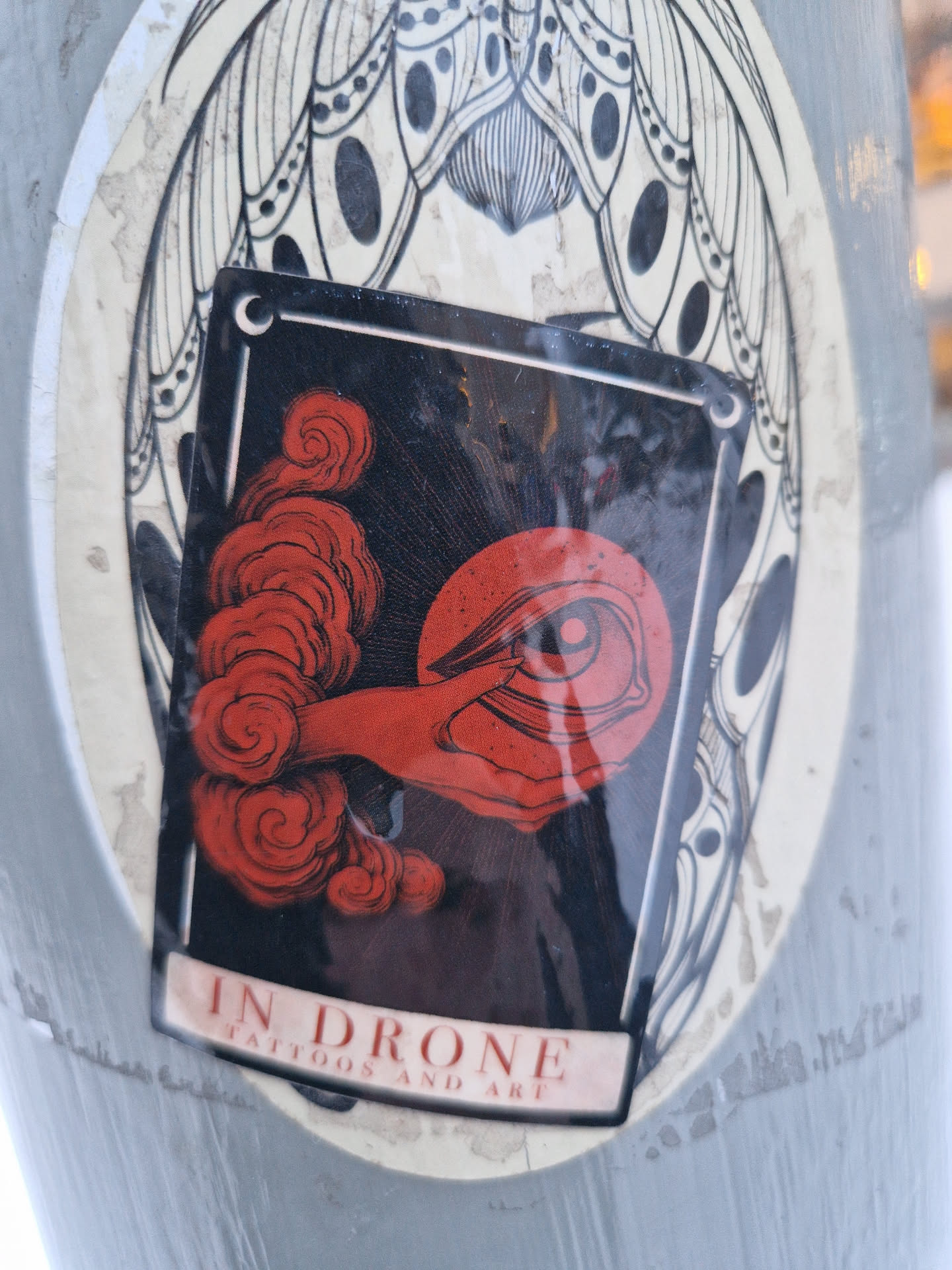A sticker featuring an illustration of a hand emerging from red clouds, holding an eye.  The style is dark and evocative, with fine line details. The sticker is affixed to a larger sticker with a moth-like design.