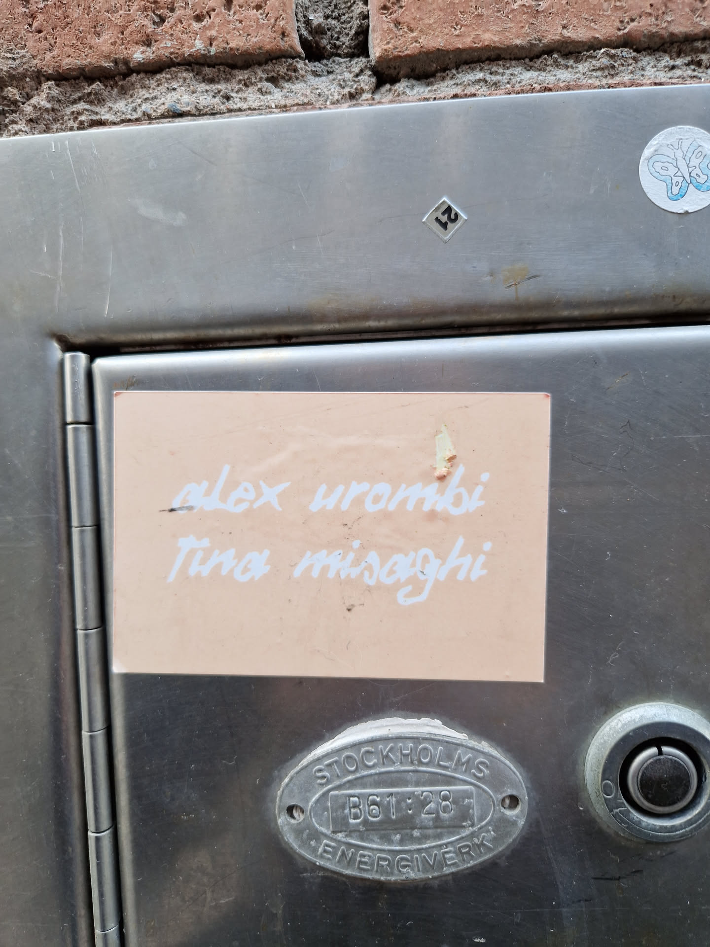 A peach-colored sticker with the names Alex Urombi and Tina Misaghi written in white. It's adhered to a metallic surface.
