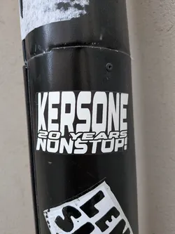 A rectangular vinyl sticker adhered to a cylindrical black metal pole. The design features bold white typography outlined in black. The top, dominant section displays the name 'KERSONE' in tall, condensed, blocky capital letters. Underneath, smaller text is stacked in two lines reading '20 YEARS' and 'NONSTOP!' in an angular, modern sans-serif typeface.