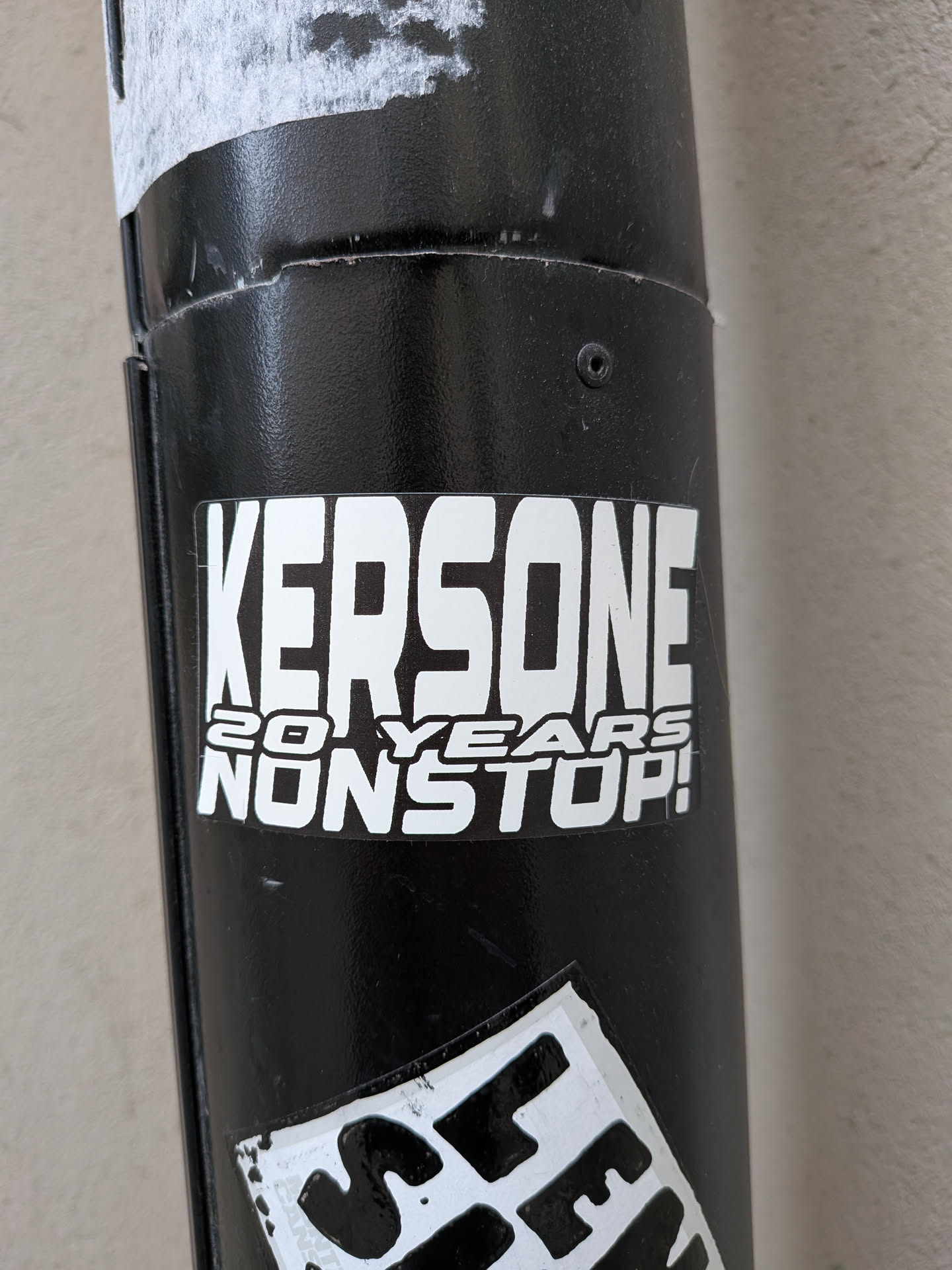 A rectangular vinyl sticker adhered to a cylindrical black metal pole. The design features bold white typography outlined in black. The top, dominant section displays the name 'KERSONE' in tall, condensed, blocky capital letters. Underneath, smaller text is stacked in two lines reading '20 YEARS' and 'NONSTOP!' in an angular, modern sans-serif typeface.
