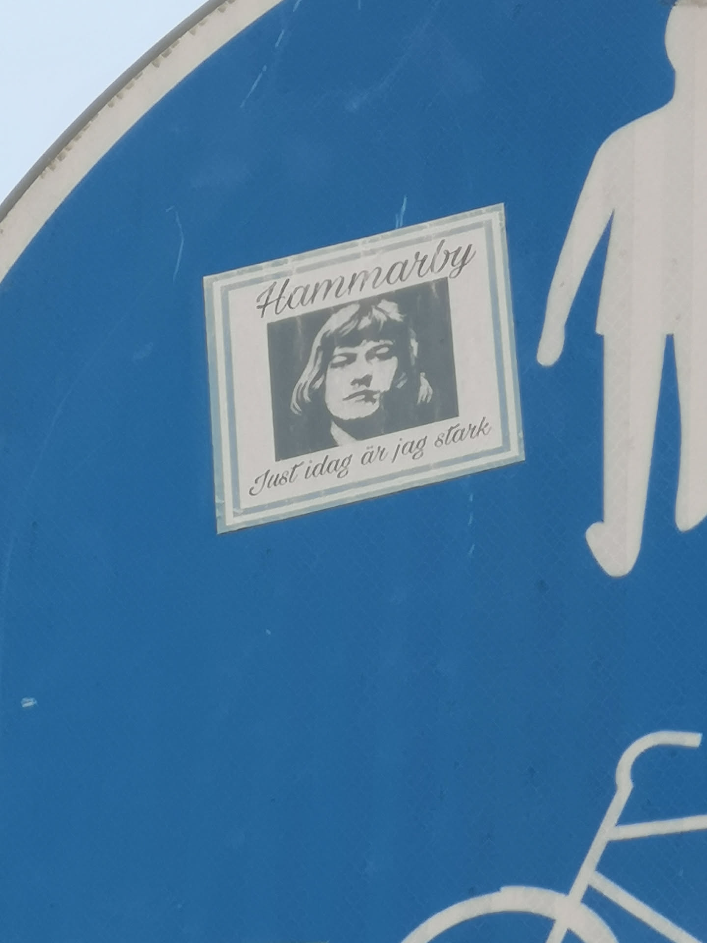 A rectangular sticker featuring a black and white (grayscale) portrait of a person, likely male, with shoulder-length hair and a contemplative expression. The portrait is framed by a light-colored double-line border. Above the portrait, in a decorative cursive font, is the word 'Hammarby'. Below the portrait, in a similar cursive font, is the phrase 'Just idag är jag stark'. The sticker has a slightly worn appearance and is affixed to a blue surface, likely a road sign, which also shows parts of a white pedestrian and bicycle symbol.