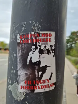 A sticker featuring a black and white image of a man, possibly protesting or demonstrating.  Red text at the top and bottom. The text translates from Danish to: "The fight against oppression is not a crime."