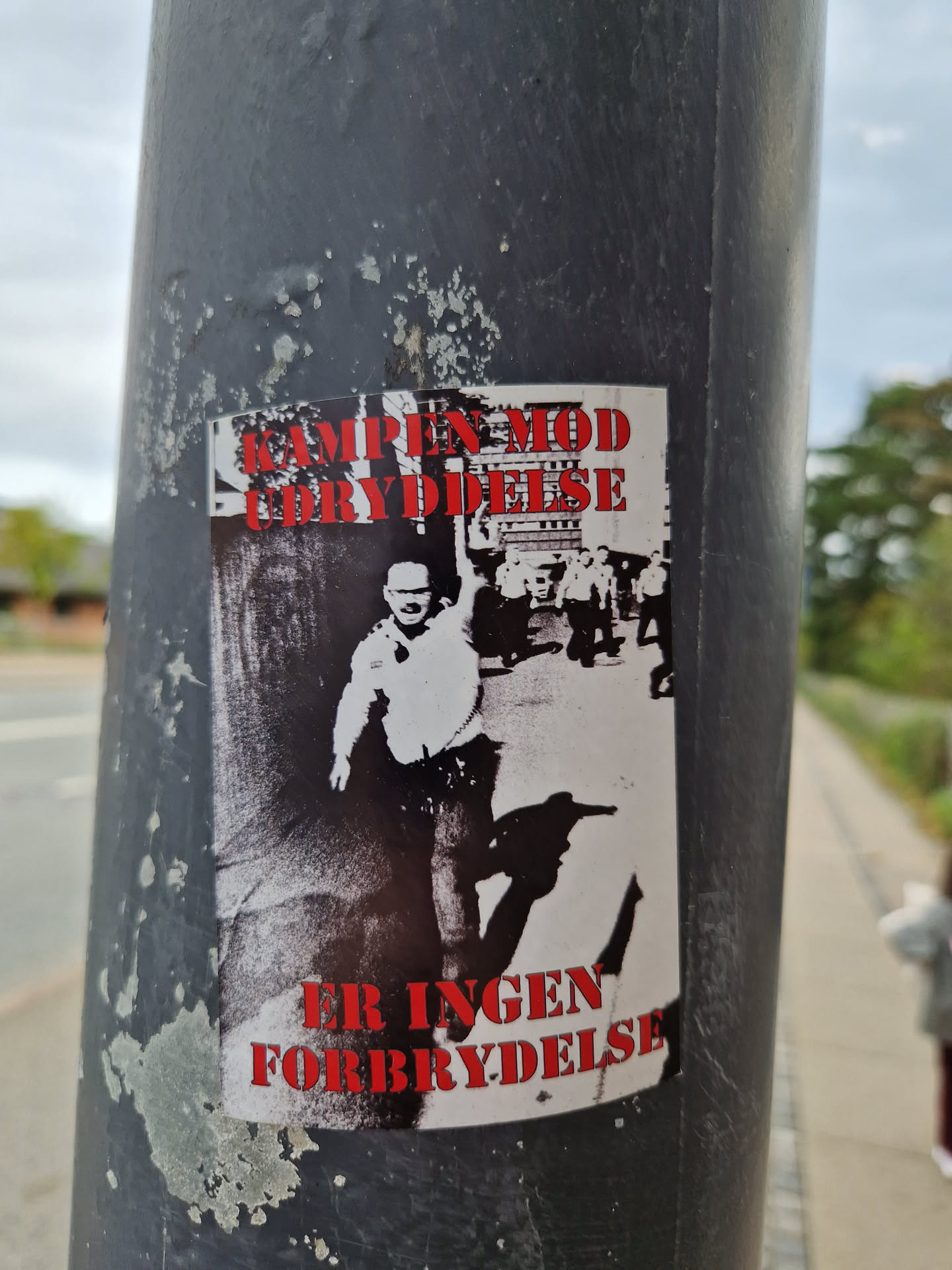 A sticker featuring a black and white image of a man, possibly protesting or demonstrating.  Red text at the top and bottom. The text translates from Danish to: "The fight against oppression is not a crime."