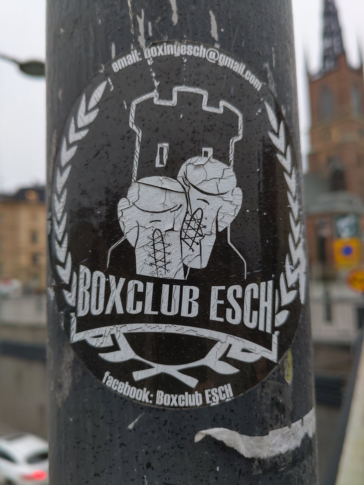 A black and white sticker with the logo of Boxclub Esch. The logo features two boxing gloves in front of a castle and has text that says "Boxclub Esch" at the bottom.  There is additional contact information on the sticker including an email and Facebook page.
