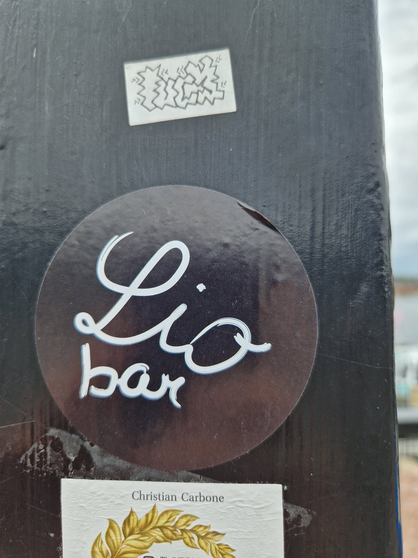 A round, dark-colored sticker with the text "Lio bar" written in a stylized, handwritten white script.  The sticker is affixed to a dark surface. There is a smaller sticker below, which appears to indicate the artist is Christian Carbone.