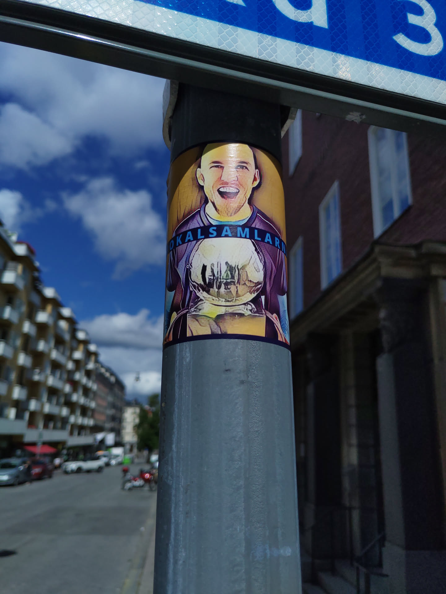 A sticker featuring a portrait-style illustration of a man with a joyful expression, seemingly holding a trophy or award. The background is a muted gold/brown tone.  The text at the bottom of the image is partially visible, suggesting possibly a name or location, but it's not fully legible. The sticker is affixed to a grey metal pole on a city street.