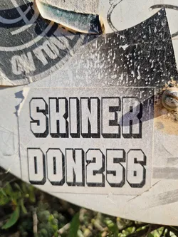 Close-up of a worn sticker with the text "SKINNER DON256" in a bold, sans-serif font. The sticker is affixed to a surface, possibly outdoors, and shows signs of wear and tear.