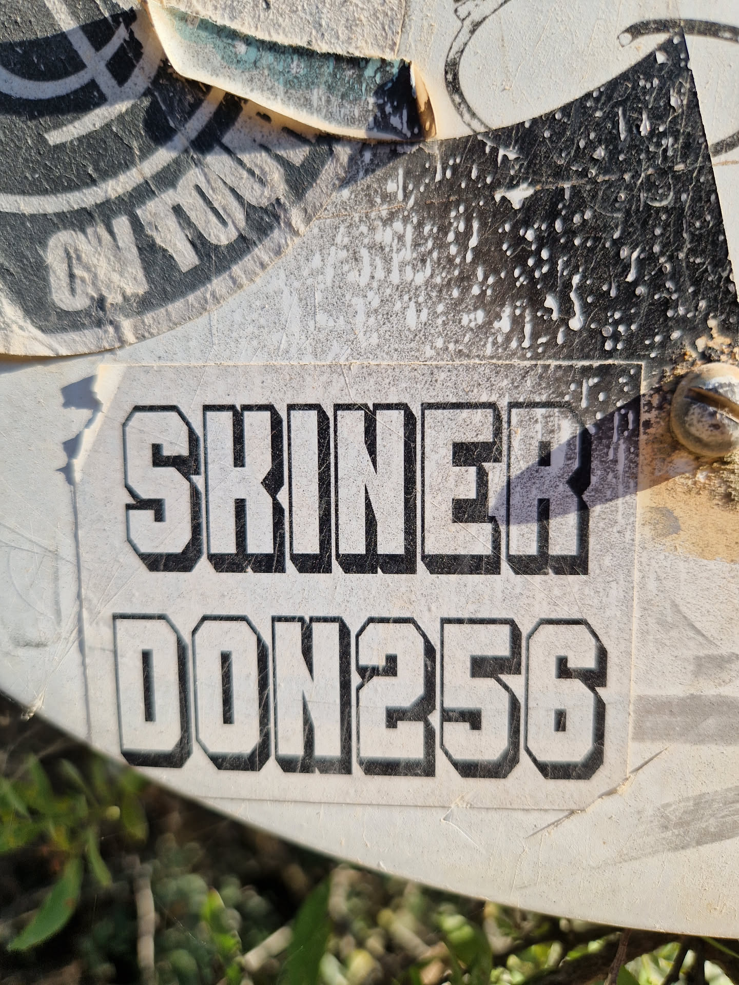 Close-up of a worn sticker with the text "SKINNER DON256" in a bold, sans-serif font. The sticker is affixed to a surface, possibly outdoors, and shows signs of wear and tear.