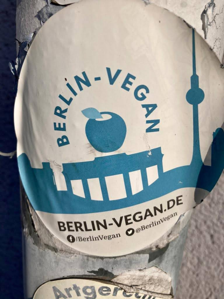 Round sticker with the text BERLIN-VEGAN in a circle around a blue apple with a leaf on top and the Berlin skyline. Contains social media links to Facebook and Twitter.