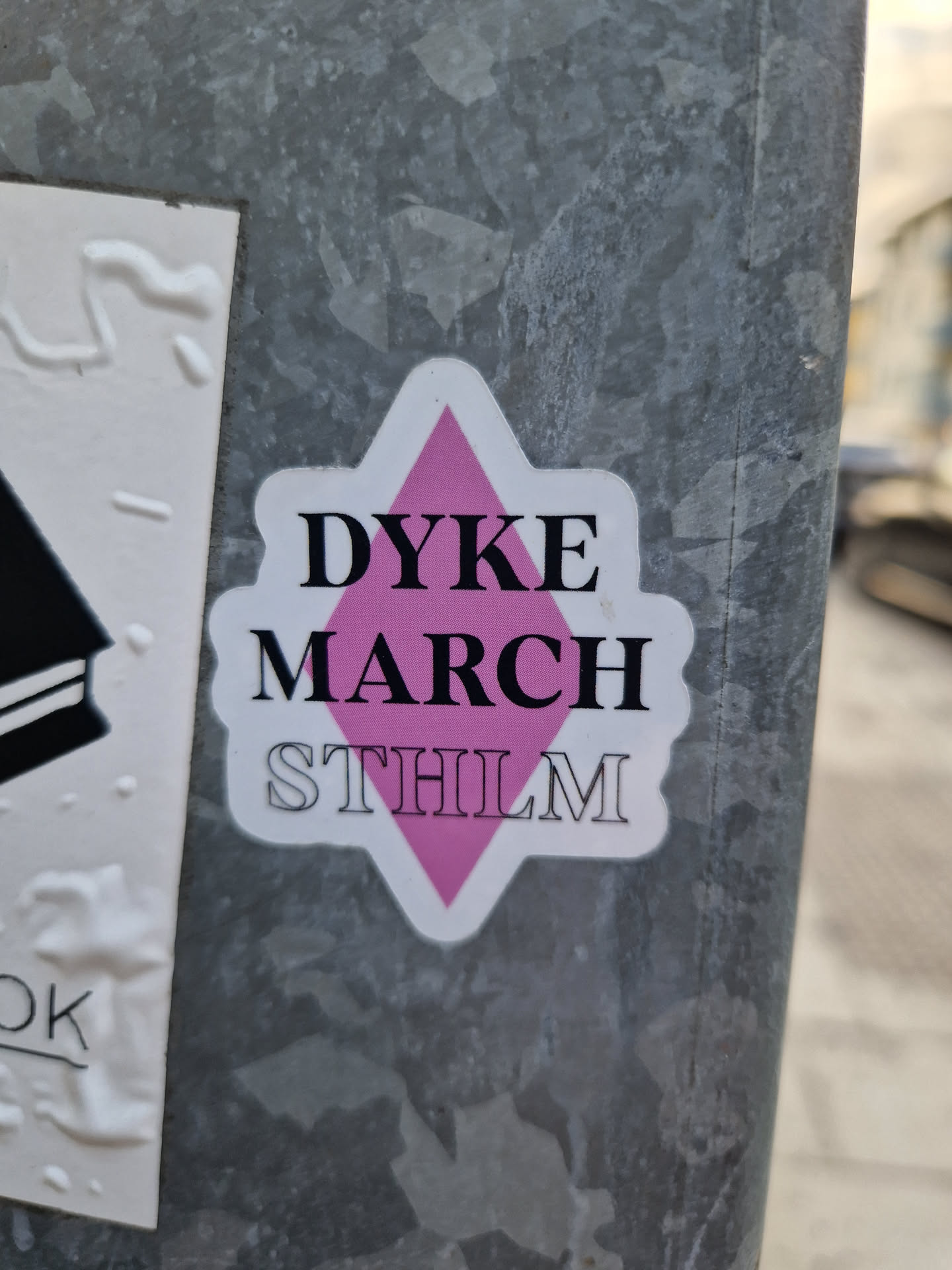 A pink diamond-shaped sticker with the text "DYKE MARCH STHLM" on it. The sticker is placed on a gray metal surface.