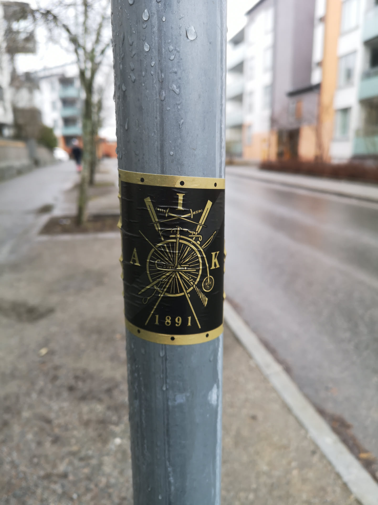 A rectangular sticker with a black background and gold-colored design, wrapped around a wet, grey metal pole. The central motif is a circular emblem featuring a detailed gold outline of a penny-farthing bicycle. Behind the bicycle, two swords are crossed. The emblem is framed by a thin gold border at the top and bottom, adorned with small black dots resembling rivets. Partial gold text is visible around the central design: 'A' on the left, 'K' on the right, and '1891' clearly at the bottom. The sticker shows signs of wear and moisture, with visible wrinkles and water droplets.