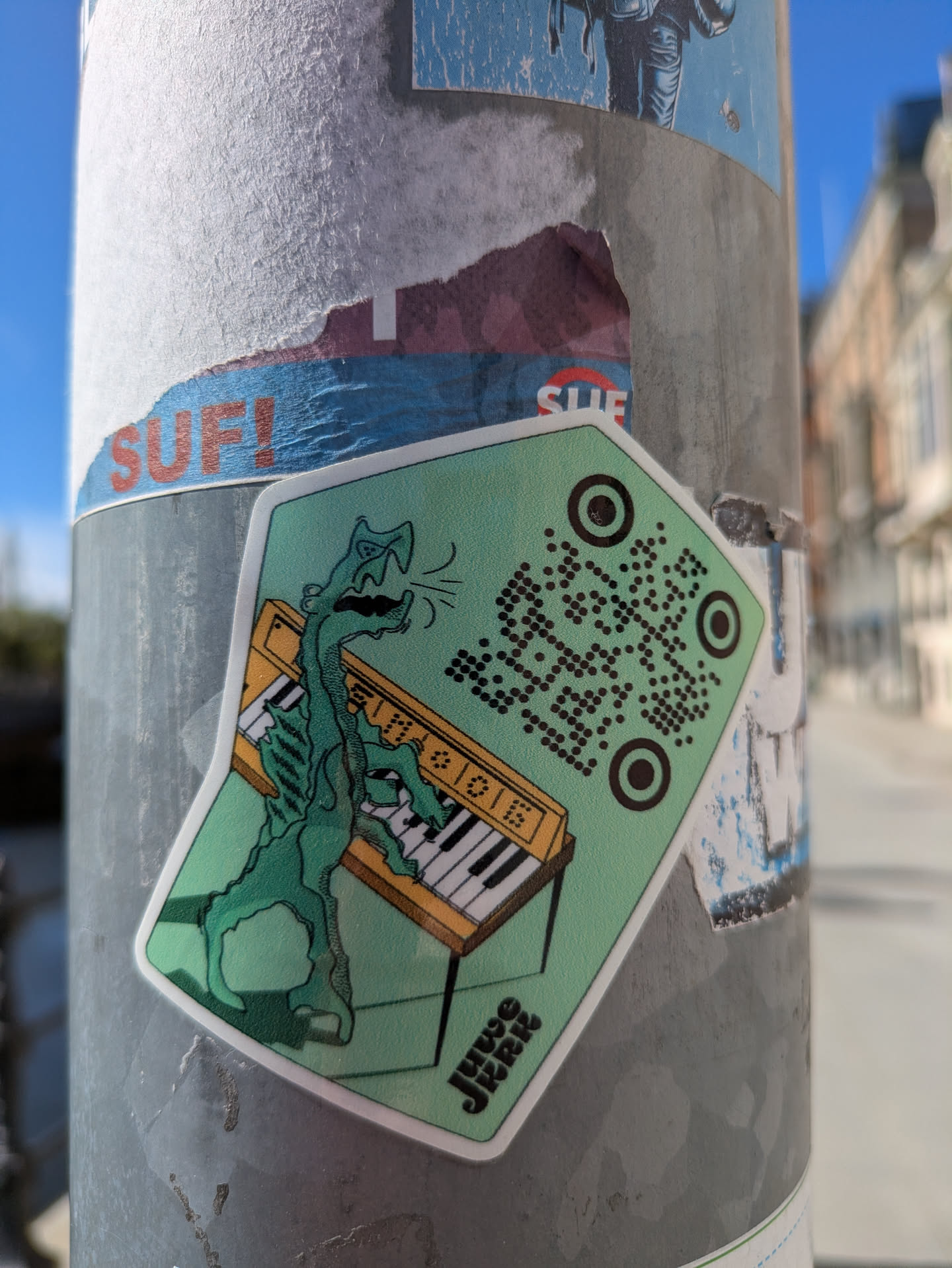 A light-green, pentagon-shaped sticker on a pole features a stylized illustration of a tall, slender, green monster with a scaly texture playing a tan-colored piano. The piano has black and white keys. The sticker includes a QR code on the top right. The name 'JUWO JRRR' is printed in stylized font at the bottom of the sticker.
