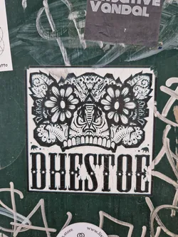 Black and white sticker with a detailed illustration of a stylized face, possibly a skull with floral elements. The word DHEDTOE is written in a bold, serif font below the illustration.