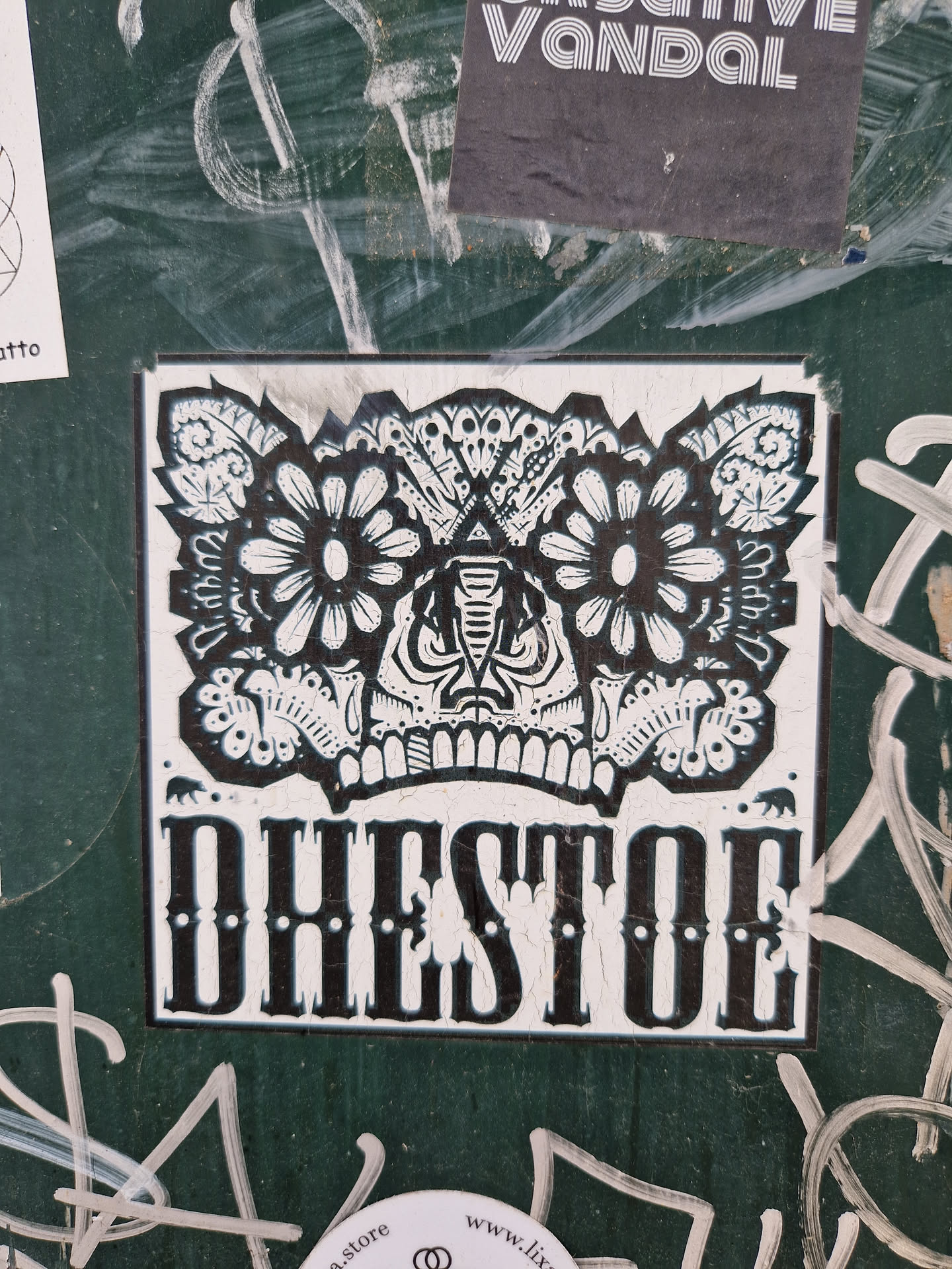 Black and white sticker with a detailed illustration of a stylized face, possibly a skull with floral elements. The word DHEDTOE is written in a bold, serif font below the illustration.