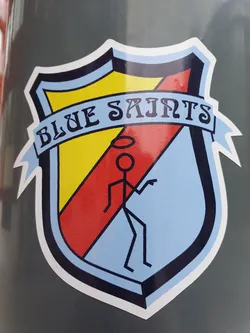 A sticker featuring a shield with the words "BLUE SAINTS" in a banner at the top. Inside the shield is a simple line drawing of a figure with a halo. The shield is colored in yellow, red and light blue.