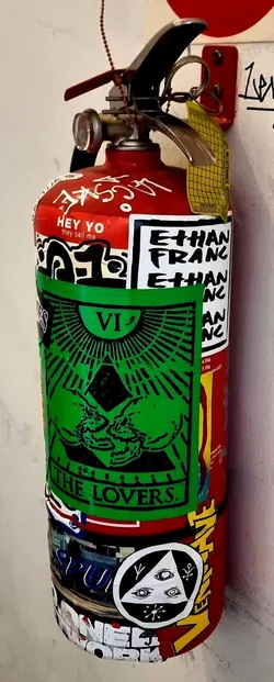 Street sticker A vertical rectangular sticker printed in black on vivid green paper, designed in the style of a traditional tarot card. At the top, the Roman numeral 'VI' sits beneath a radiating semi-circle. In the center, two stylized human skulls lean close together in front of a dark geometric diamond, encircled by radiating beams. The bottom is bordered and inscribed with the bold serif text 'THE LOVERS.'