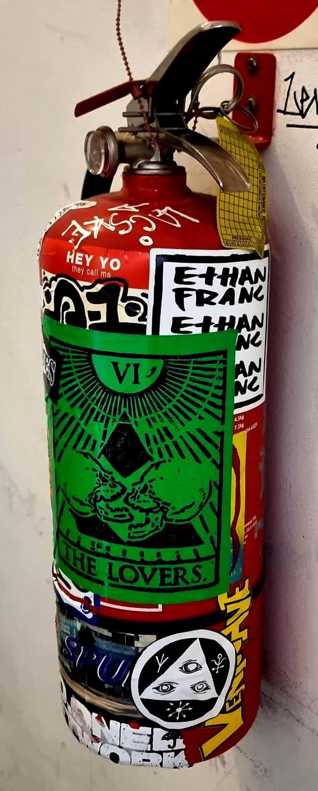 A vertical rectangular sticker printed in black on vivid green paper, designed in the style of a traditional tarot card. At the top, the Roman numeral 'VI' sits beneath a radiating semi-circle. In the center, two stylized human skulls lean close together in front of a dark geometric diamond, encircled by radiating beams. The bottom is bordered and inscribed with the bold serif text 'THE LOVERS.'