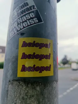 A yellow sticker with the word 'heiopei' repeated three times in a stylized purple font.  The sticker is affixed to a gray pole, likely outdoors.  The meaning of 'heiopei' is currently unknown. There are no social media links or QR codes visible on this sticker.