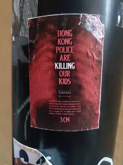 A sticker with a dark red background and white text that reads "HONG KONG POLICE ARE KILLING OUR KIDS". There is also some smaller text at the bottom that describes an incident. The sticker is attached to a dark-colored surface.