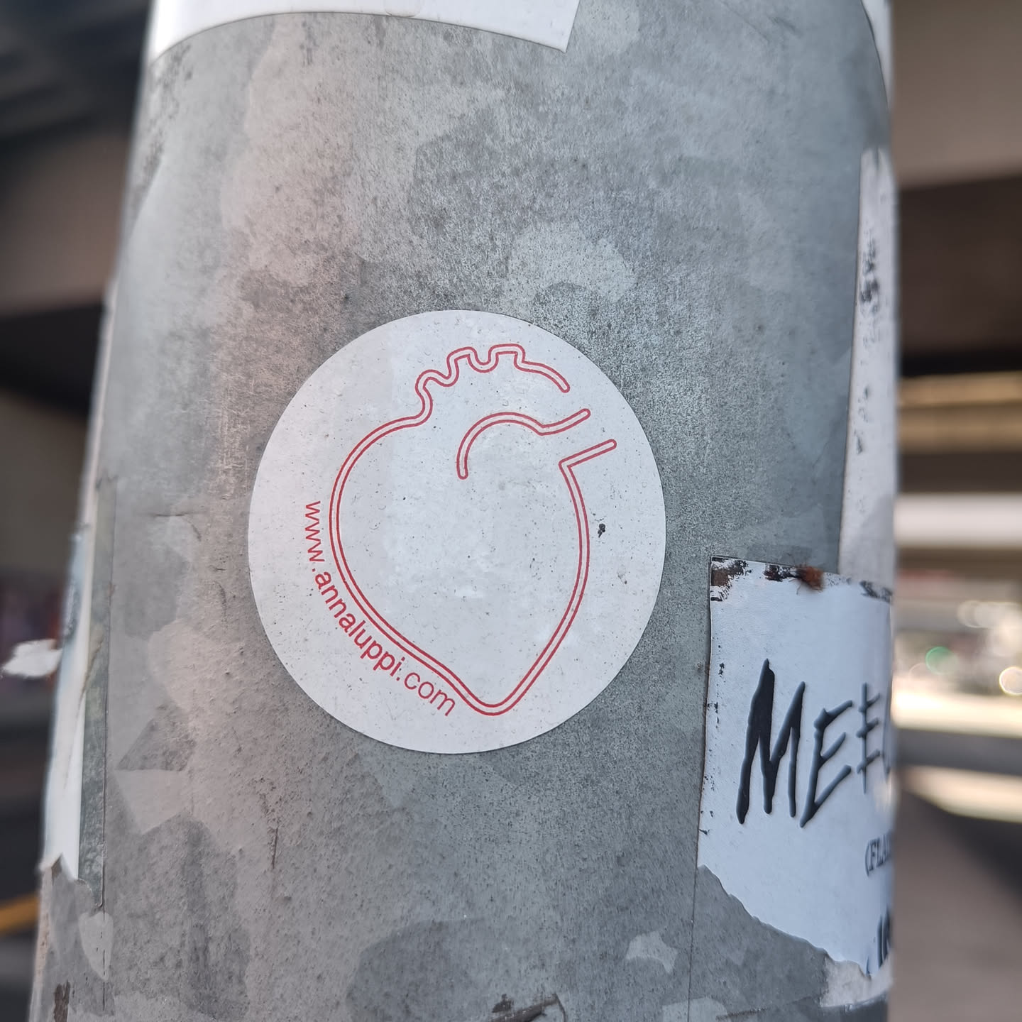 A white circular sticker with a red line drawing of a heart. The website address www.annaluppi.com is printed in small red text beneath the illustration.