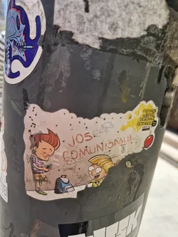 Sticker featuring an illustration of two cartoon children, a boy and a girl, with graffiti-style text in the background that says "JOS Comunisra!!". The boy is listening to music on headphones and holding a small device. The girl appears to be smiling. There's a backpack on the ground next to them.