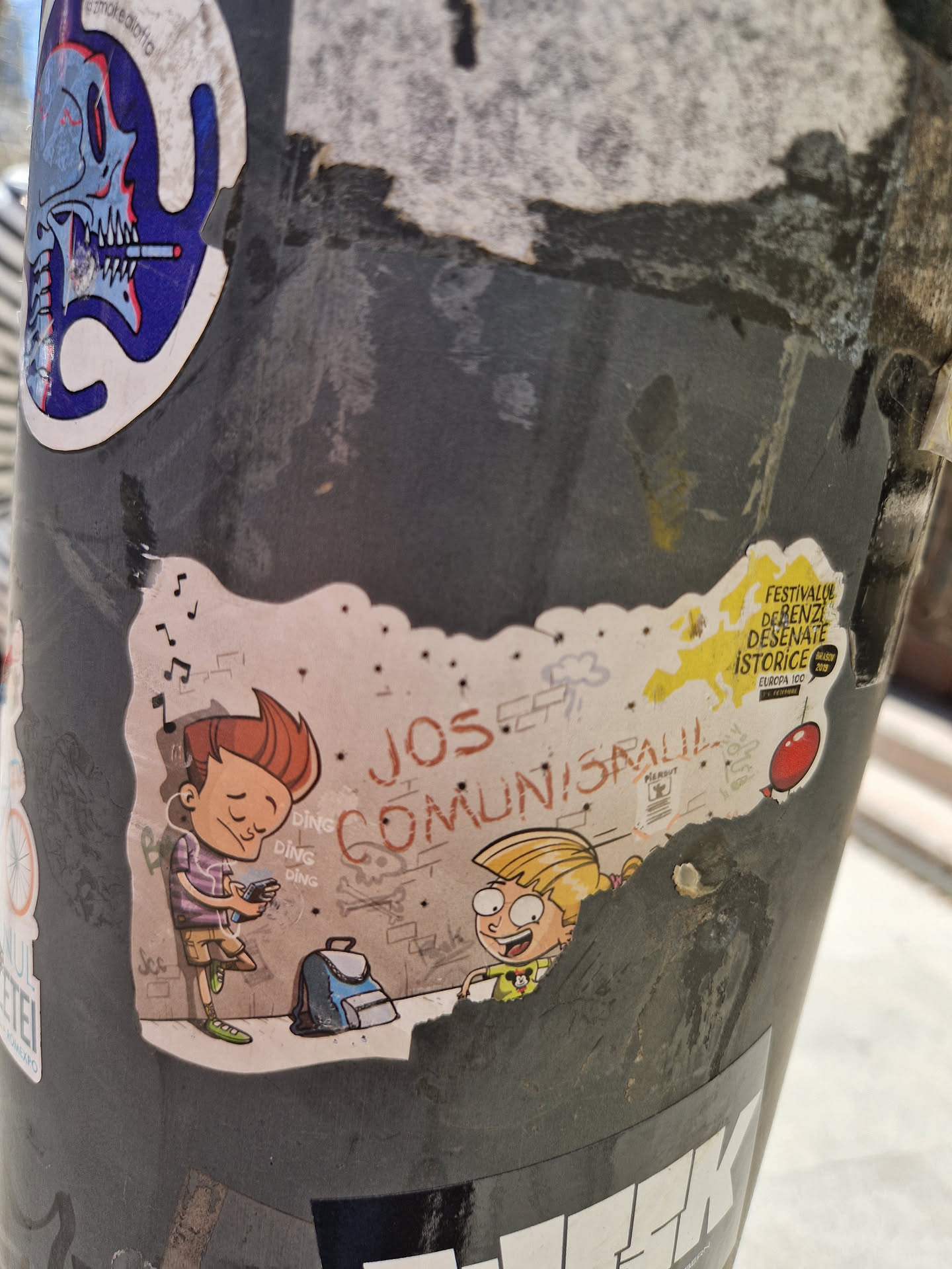 Sticker featuring an illustration of two cartoon children, a boy and a girl, with graffiti-style text in the background that says "JOS Comunisra!!". The boy is listening to music on headphones and holding a small device. The girl appears to be smiling. There's a backpack on the ground next to them.