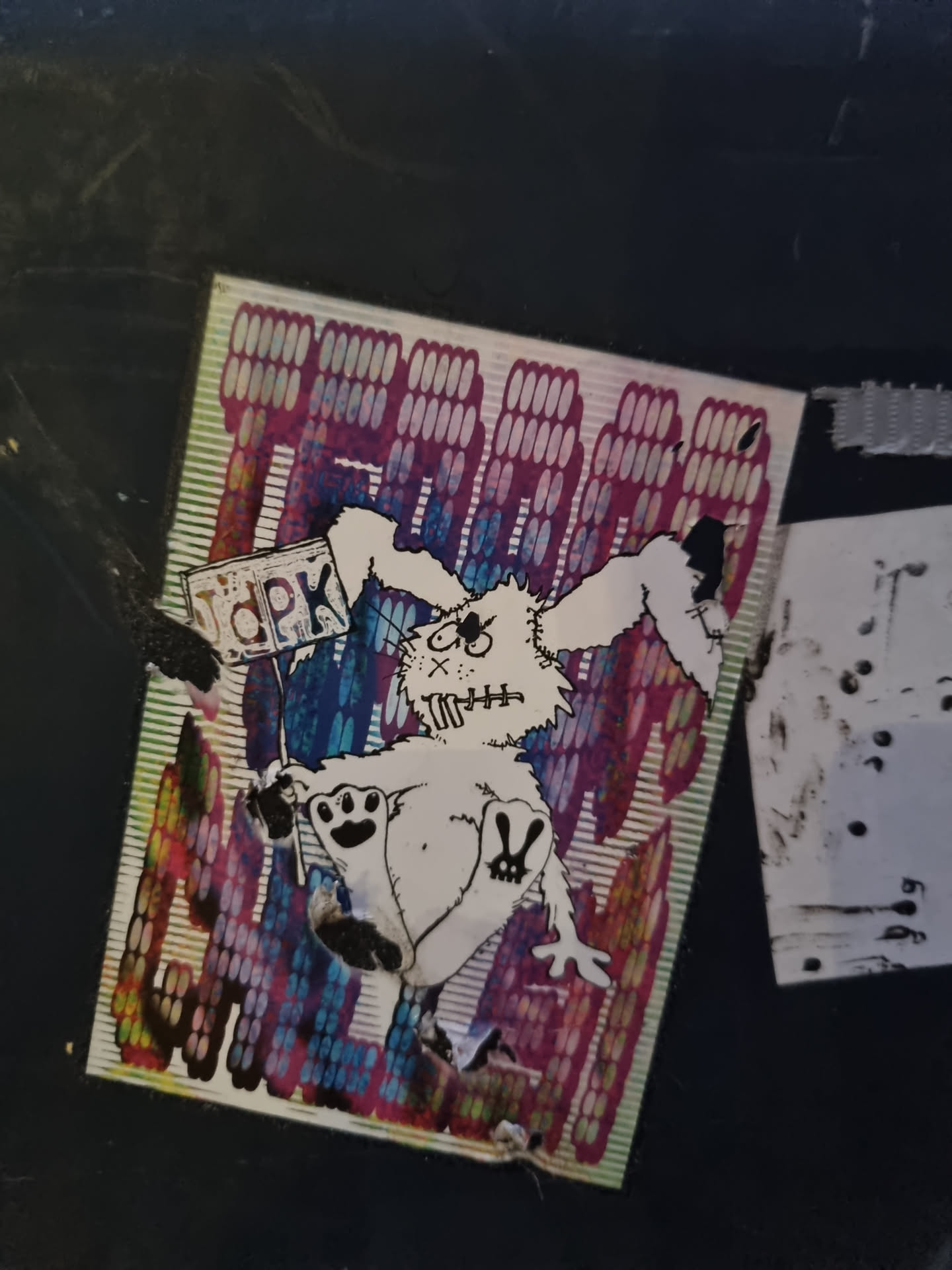 This sticker features a cartoon-like bunny character with stitched-up features, holding a sign that seems to say 'TPK'. The bunny is depicted in a playful, almost mischievous way.  The background has a repeating pattern in vibrant, multicolored vertical stripes, creating a dynamic and eye-catching effect. The sticker is slightly worn, suggesting it has been placed outdoors.