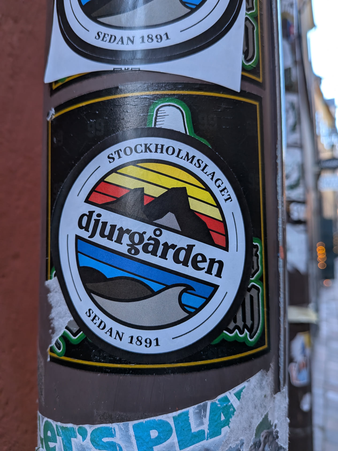 The sticker is circular, featuring a prominent white outer ring with a black background behind the central emblem. The main circular emblem is divided horizontally into two distinct sections. The upper section depicts a stylized sunset or sunrise with horizontal stripes in yellow, orange, and red, overlaid with dark grey or black silhouettes of mountains. The lower section features a blue sky or water, a darker blue stripe, and a wavy grey-brown foreground representing land or water. Text 'STOCKHOLMSLAGET' is curved along the top of the white ring, 'djurgården' is prominently displayed in black lowercase letters across the center, and 'SEDAN 1891' is curved along the bottom of the white ring. The overall design evokes a vintage or retro aesthetic, possibly a crest or logo for a sports club or local identity.