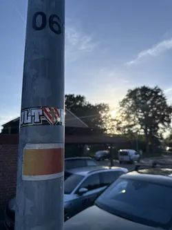 Street sticker A rectangular sticker wrapped around a grey metal utility pole. The sticker features the bold white text 'ULT' with a black outline on the left, followed by a central shield emblem. The shield is divided diagonally into red and white sections and features a gold cross in the center. To the right of the shield, the sticker continues around the pole. Below it, a second, simpler sticker with a yellow-to-red gradient is also visible.
